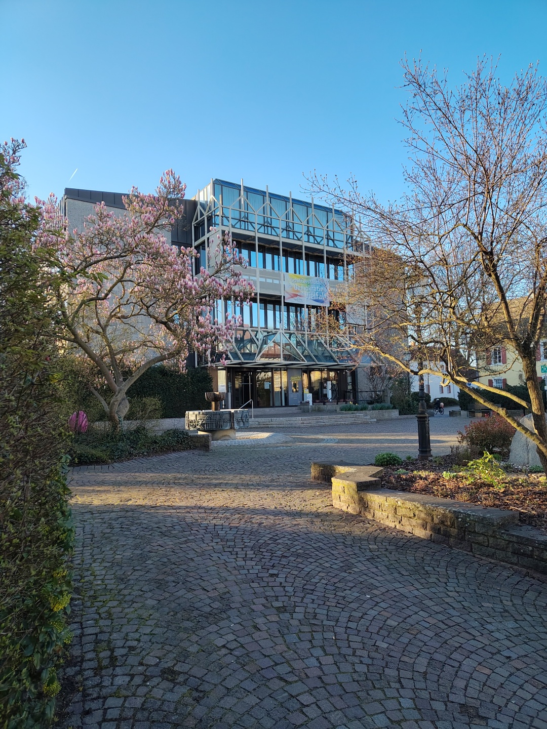 Im Vordergrund ist der blühende Magnolienbaum und der Brunnen mit einem großen Osterei zu sehen, im Hintergrund das Rathaus.