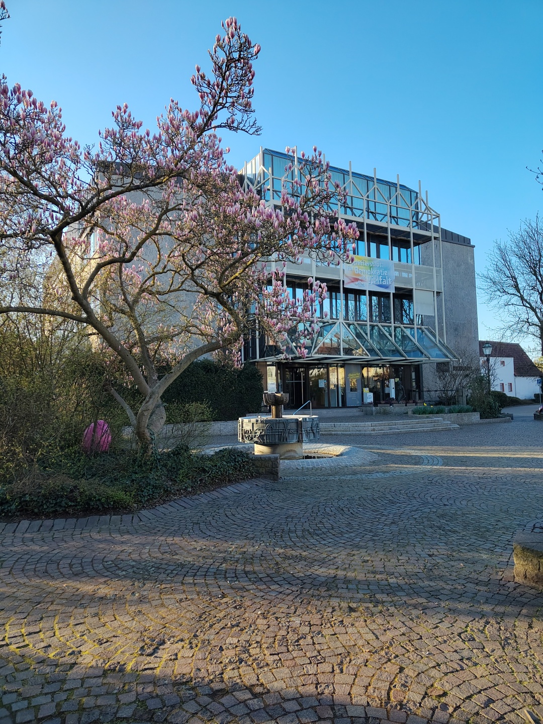 Im Vordergrund ist der blühende Magnolienbaum und der Brunnen mit einem großen Osterei zu sehen, im Hintergrund das Rathaus.