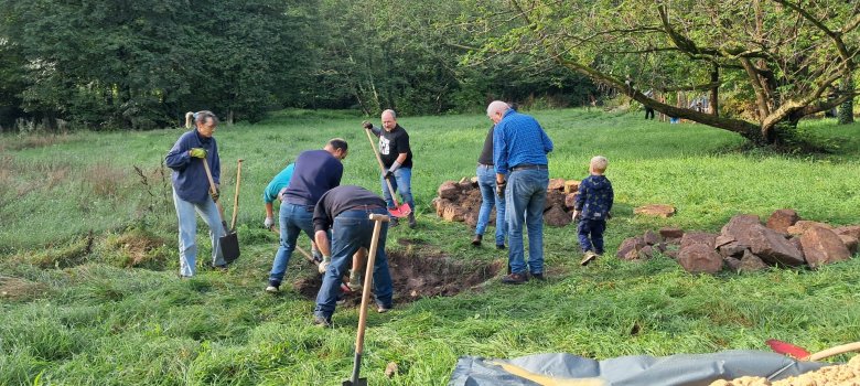 Männer, Frauen mit Schaufeln bearbeiten in einer Wiese Bereiche. Im Vordergrund liegt ein Berg Sand.