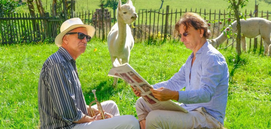 zwei Männer auf einer Wiese im Vordergrund, einer badet seine Füße, der andere liest Zeitung