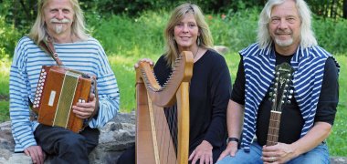 Zwei Männer und eine Frau sitzen mit Instrumenten in den Händen auf einer Mauer
