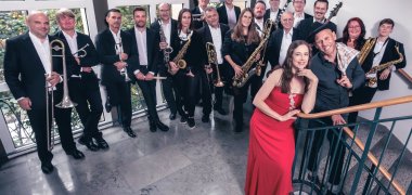 Männer und Frauen schwarz-weiß angezogen mit Instrumenten in den Händen stehen im Hintergrund. Im Vordergrund steht eine Frau in einem roten Kleid.